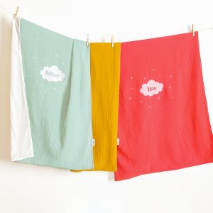 Plaids vert, jaune et rose personnalisés avec un prénom sur un motif nuage, suspendues sur une corde.