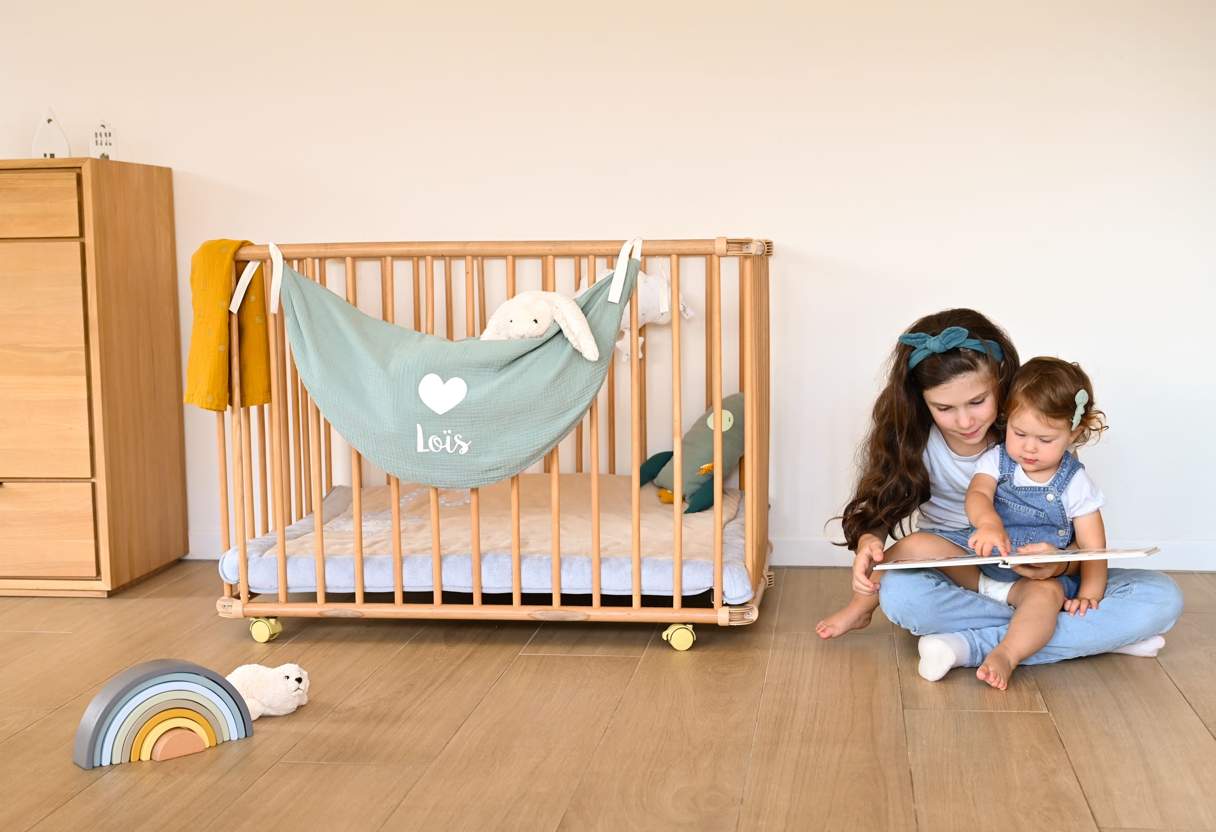 Une grande soeur lit une histoire à sa petite soeur dans une chambre joliment décorée avec un hamac à doudous.